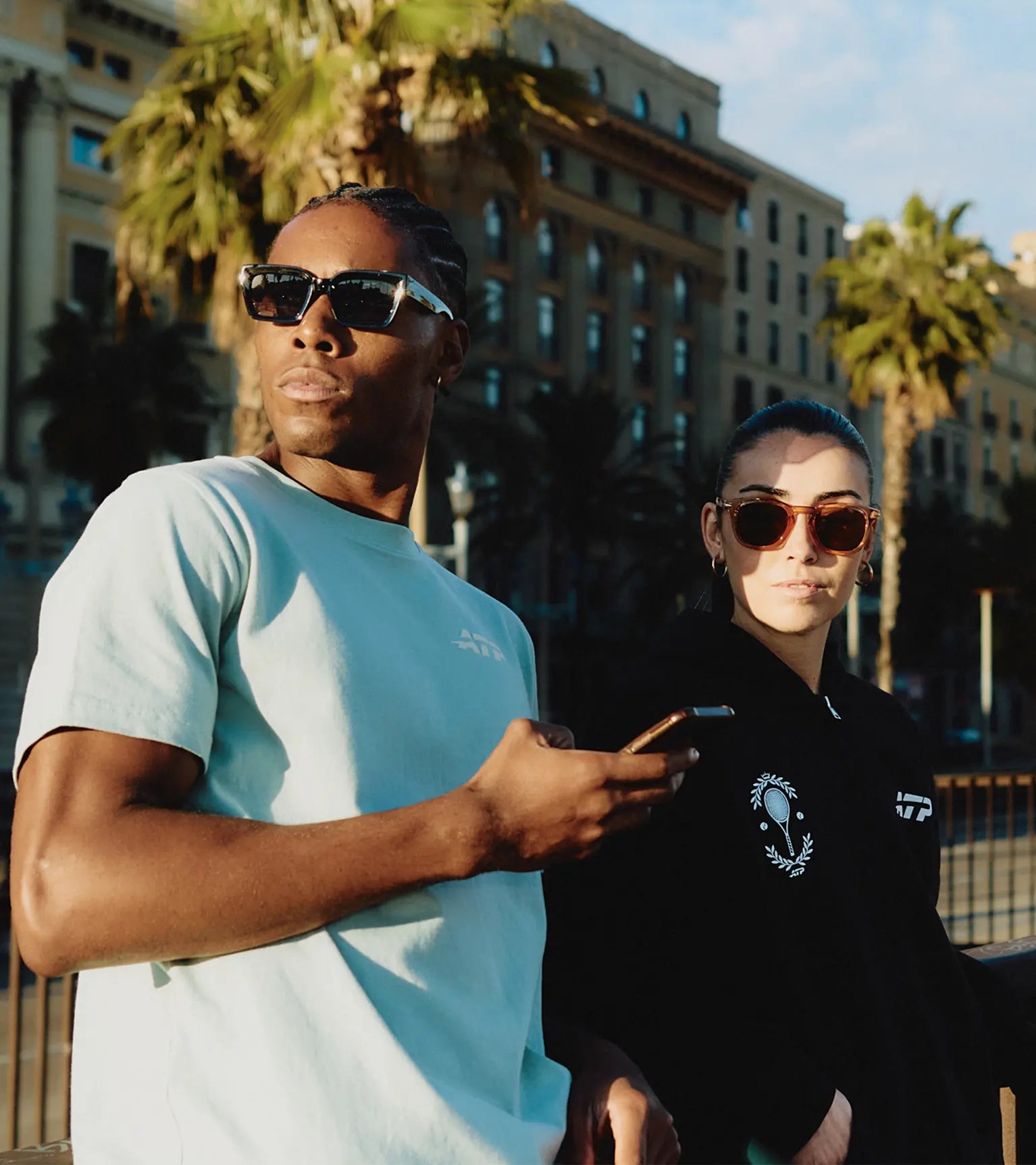 Two people wearing ATP clothing in an urban setting with palm trees and a building in the background.