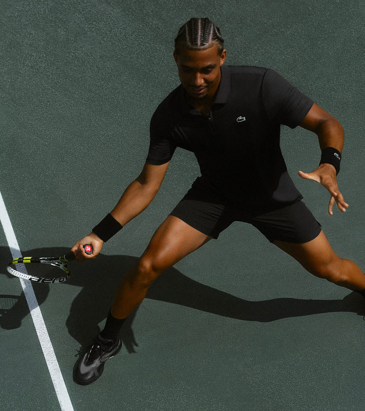 A tennis player in a black Lacoste athletic set (shirt and shorts) lunges on a green hard court, holding a racket, with a white line visible on the left.