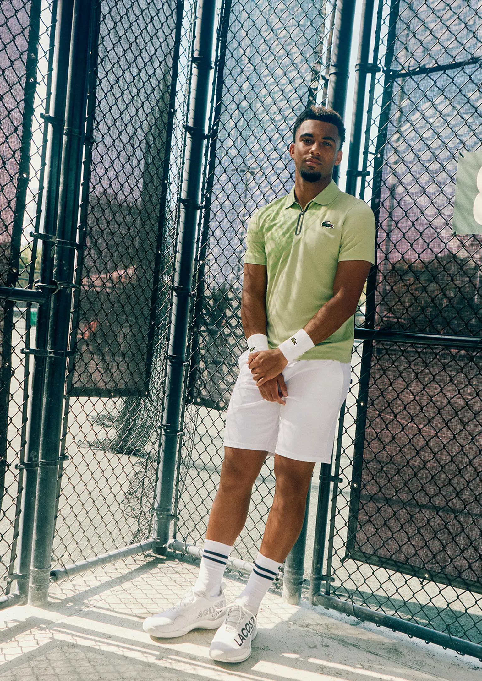 Person standing inside a fenced tennis court wearing a green Lacoste t-shirt and white shorts.